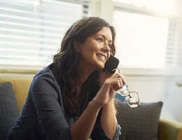 A smiling woman holds her eyeglasses and talks on a phone.