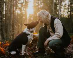 In a wooded area, a woman squats to touch noses with her dog