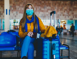 A woman wearing a mask and holding a passport sits next to a suitcase.