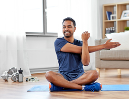 A man sits on an exercise mat and stretches his arm.