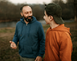 A father and his teenage son walk and talk near a pond.