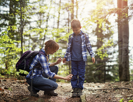 In the woods, a woman sprays a boy’s jeans with bug repellent.