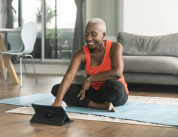 A smiling woman sits on a yoga mat and looks at a tablet.