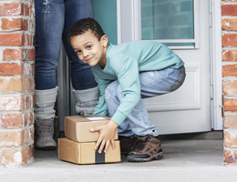A boy picks up packages on his doorstep, with his mother’s legs visible behind him.